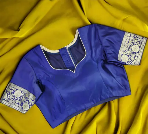 A professionally tailored royal blue silk saree padded blouse by Tailor Boutiques, featuring a sophisticated sweetheart neckline accented with delicate light golden piping. The blouse highlights expert craftsmanship with structured princess-cut stitching for a perfect fit and elbow-length sleeves adorned with intricate silver zari floral borders. Set against a contrasting draped mustard yellow fabric, this custom-stitched blouse showcases the premium finishing and boutique-quality ethnic wear design available for women in Noida.