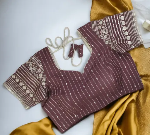Expertly stitched designer blouse in deep wine-purple featuring fine vertical sequin lines on the body and heavy floral golden zari and gota patti embroidery on the short sleeves. The blouse has a sweetheart neckline, a neat back opening, and golden thread doris (ties). This detailed image showcases the high-quality stitching and detailed embroidery work offered by Tailorboutiques.com's doorstep tailoring service in Noida Sector 76, specializing in custom women's wear like blouses, suits, and lehengas.