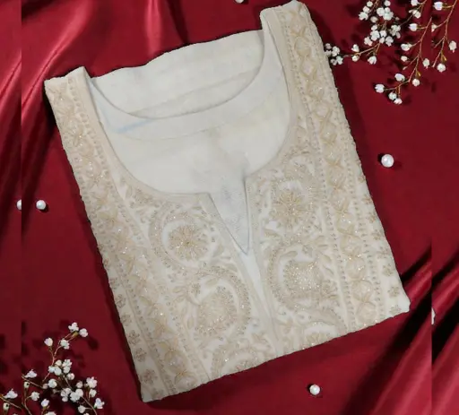 An elegantly folded off-white designer kurta featuring intricate floral thread-work and a classic V-slit neckline, resting on draped golden-yellow satin fabric with scattered white baby's breath flowers. Showcasing the premium custom stitching and bespoke ethnic wear craftsmanship offered by TailorBoutiques.com in Delhi.