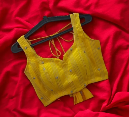 A beautifully crafted yellow sleeveless saree blouse featuring a sweetheart neckline, delicate embroidery, and a stylish pleated bottom hem, displayed on a hanger against a vibrant red backdrop. This exquisite custom-stitched garment exemplifies the premium craftsmanship and doorstep services offered by the leading ladies tailor in Delhi, Tailorboutiques.com.