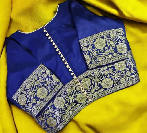 Close-up view of a stunning royal blue custom-stitched saree blouse featuring intricate silver floral zari work on the sleeves and waist border, complete with an elegant center button placket, resting beautifully on a vibrant yellow draped fabric. This exquisitely crafted ethnic garment highlights the impeccable finish, premium quality, and convenient online tailoring services offered by Tailorboutiques.com.