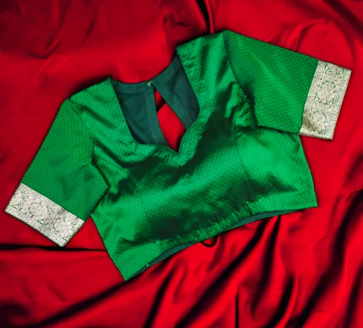 A beautifully tailored emerald green silk saree blouse featuring intricate silver zari embroidery on the sleeve cuffs and a keyhole back design, resting on a draped red silk fabric background. This example of high-quality custom tailoring is featured on the Tailoring Blog by Tailor Boutiques.